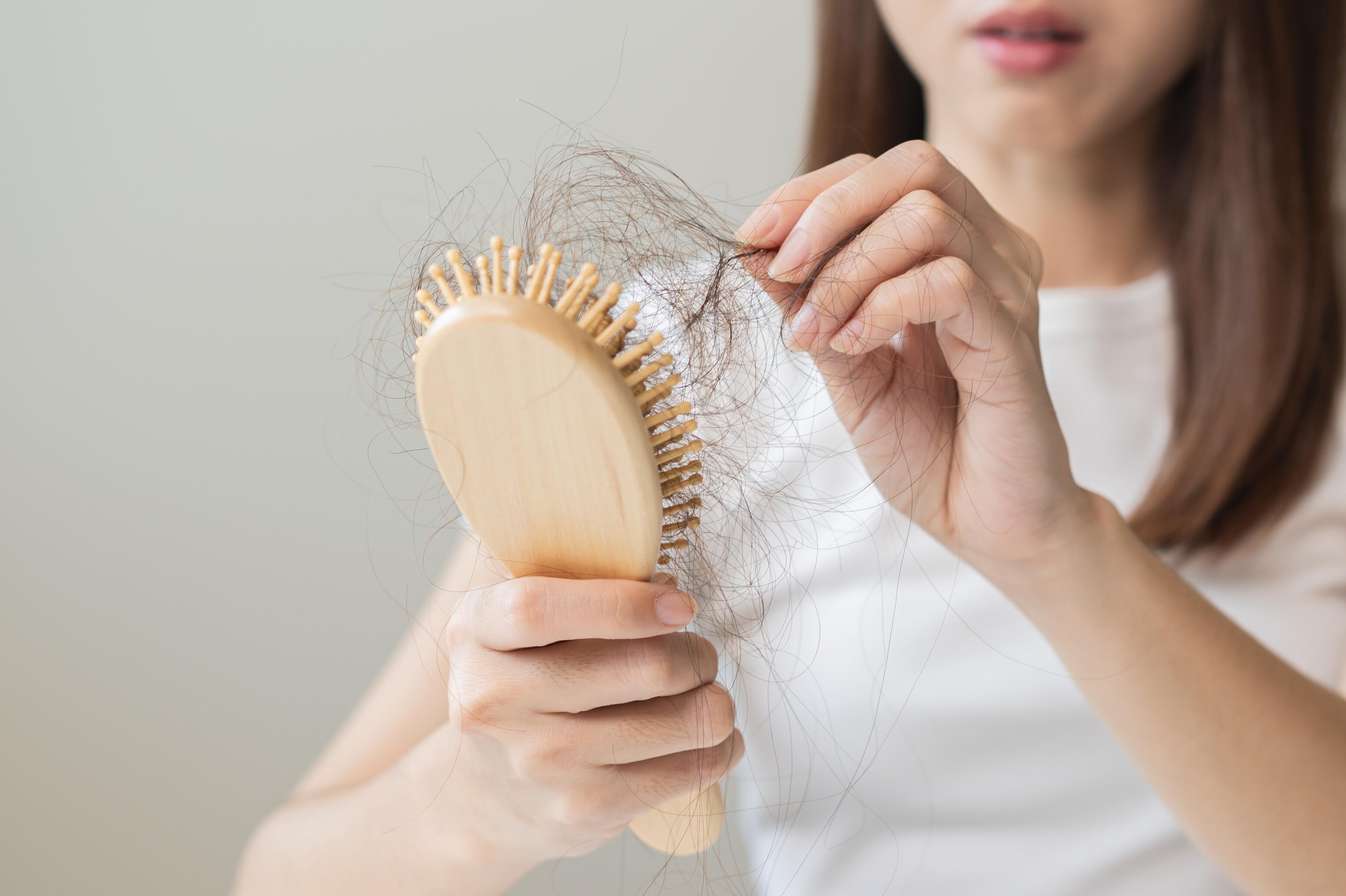 Eine dunkelhaarige Frau in einem weißen T-Shirt hält eine hölzerne Haarbürste und zieht viele lose Haare aus der Bürste.